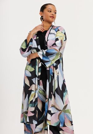 Femme portant une longue veste à motifs floraux colorés et un pantalon assorti sur un haut noir, posant avec la main près du menton.