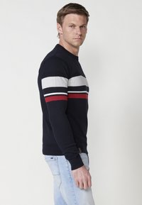 Pulover tricotat bleumarin cu dungi orizontale în gri, alb și roșu. Design ajustat, manșete și tiv cu dungi. Detaliu subtil cu logo în partea de jos.
