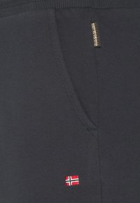 Zwarte stof met een zijzak, een kleine geborduurde Noorse vlag en een verticale Napapijri merketiket vlak bij de tailleband.