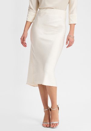 Cremfarbene Satin-Midikleid mit subtilen Punktmuster, figurbetonter Silhouette und seitlichem Schlitz; kombiniert mit silbernen Riemchensandalen.