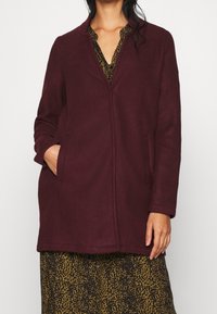 Bordeauxrode wollen jas met een lichte glans, zonder kraag, zijzakken en een rechte snit, gedragen over een jurk met patroon.