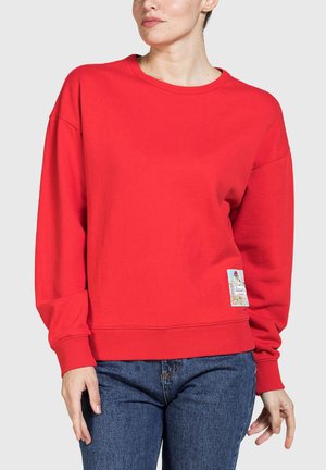 Donna che indossa una felpa rossa brillante a maniche lunghe con scollo tondo e una piccola toppa colorata nella parte inferiore sinistra, abbinata a jeans blu.