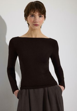 Mujer con cabello corto y castaño, vestida con una blusa negra ajustada de manga larga y pantalones plisados gris oscuro, de pie con las manos en los bolsillos sobre un fondo blanco.