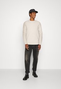 Suéter de malha bege, jeans escuros e botas de tornozelo pretas; modelo usa um chapéu escuro. Design simples, textura suave, ajuste descontraído.