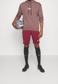Sweat à capuche marron avec logo, shorts marron, chaussettes noires montantes jusqu'aux genoux, et chaussures de football noires ; tenant un ballon de football Nike gris.
