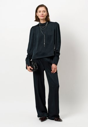 Zwarte, gestructureerde blouse met lange mouwen en een hoge hals, gecombineerd met losvallende zwarte broek. Accessoires zijn onder andere gelaagde zilveren kettingen en een clutch.