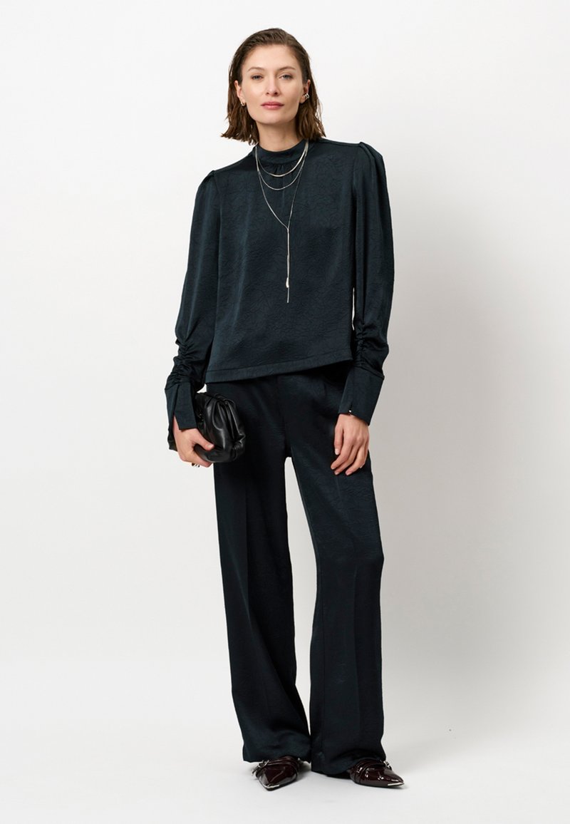 Schwarze, strukturierte Bluse mit langen Ärmeln und hohem Kragen, kombiniert mit lockeren schwarzen Hosen. Als Accessoires dienen übereinander getragene silberne Halsketten und eine Clutch.