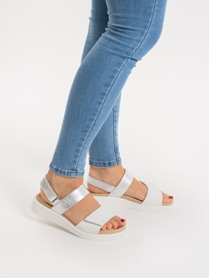 Füße, die silber-weiße Keilsandalen tragen, kombiniert mit blauen Skinny-Jeans, die rot lackierte Zehennägel auf weißem Hintergrund zeigen.