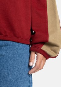 Rode sweatshirt met beige mouwen, ribgebreide boorden en zoom. Draagt over blauwe jeans, de stof lijkt zacht en textuur te hebben.
