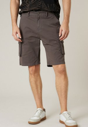 Homme portant un short cargo gris foncé avec des poches latérales à boutons et des baskets blanches, debout devant un fond uni.