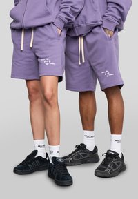Lichtpaarse shorts met beige trekkoord en logodetail, gecombineerd met zwarte sneakers en witte sokken, getoond op twee personen.