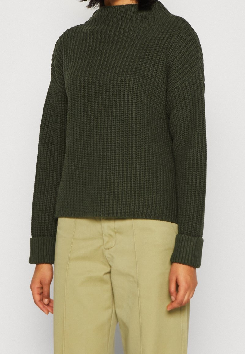 Pull en tricot vert foncé avec un col haut, des manches longues et une texture côtelée. Associé à un pantalon beige clair.