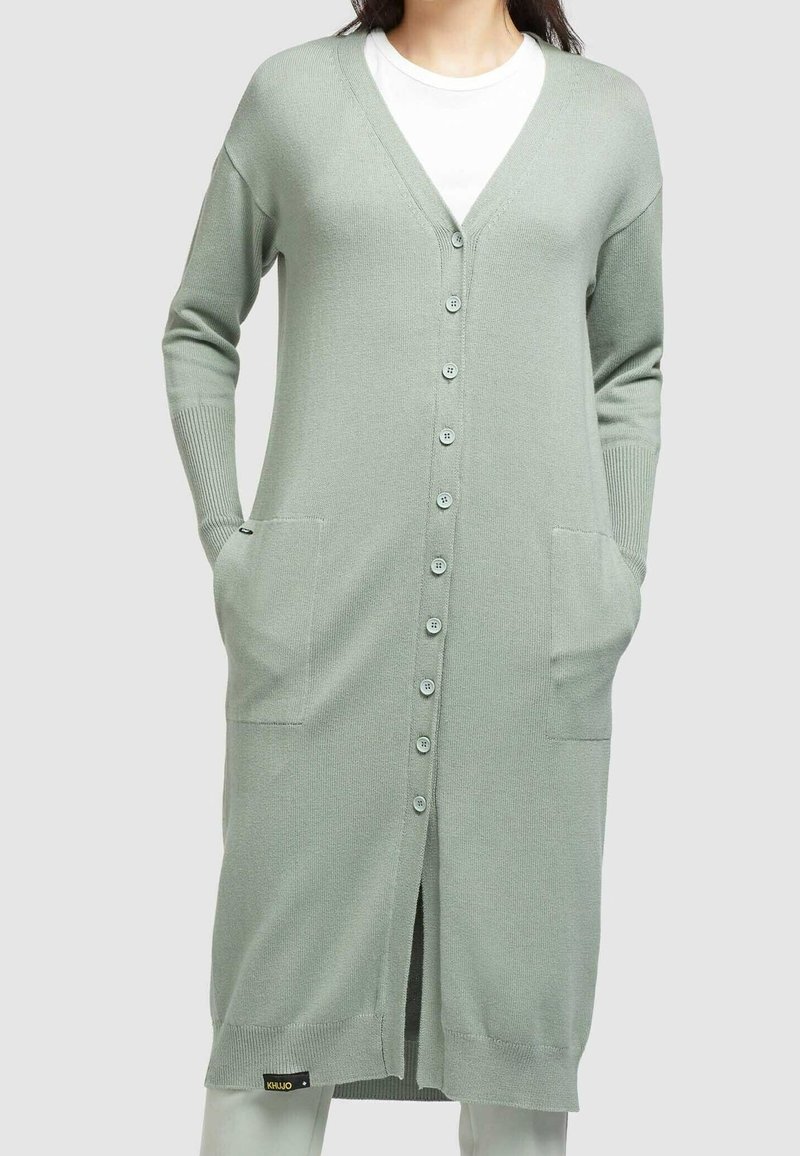 Femme portant un long cardigan vert clair boutonné avec des poches par-dessus une chemise blanche, debout, les mains dans les poches.
