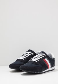 Sneakers z czarną siateczką, zamszowymi akcentami, białymi sznurówkami i czerwonymi paskami. Posiadają białą gumową podeszwę z teksturowanym wzorem zapewniającym przyczepność.