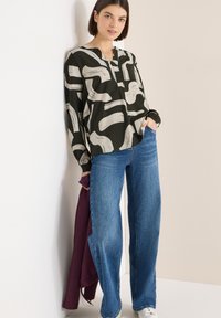 Černá halenka s bílým abstraktním vzorem štětcových tahů, zapnutý výstřih na knoflíky, v kombinaci s modrými širokými džíny a vínově červenou bundou přehozenou přes jedno rameno.