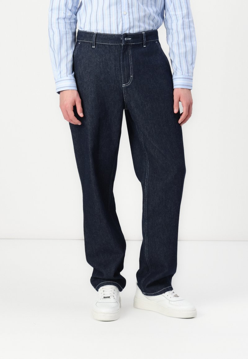 Mörk denimbyxor, rak passform, med kontrasterande vit sömnad och knäppning i midjan. Bärs tillsammans med en ljusblå randig skjorta och vita sneakers.