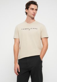 Μπεζ βαμβακερό μπλουζάκι με λογότυπο "TOMMY JEANS" σε μαύρο χρώμα, στρογγυλή λαιμόκοψη και κοντά μανίκια, συνδυασμένο με μαύρο παντελόνι.