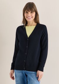 Femme souriante portant un cardigan noir boutonné par-dessus un haut jaune et un jean bleu, debout devant un fond clair uni.