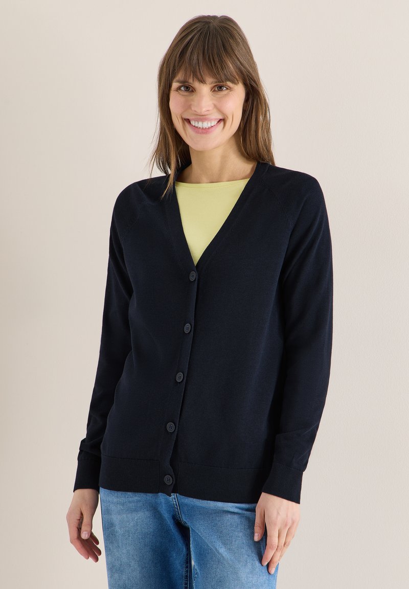 Femme souriante portant un cardigan noir boutonné par-dessus un haut jaune et un jean bleu, debout devant un fond clair uni.