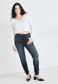 Λευκή κοντή μπλούζα με μακριά μανίκια και σκούρες μπλε στενές τζιν. Το μοντέλο φοράει μαύρα τακούνια με μυτερή μύτη. Λεπτό ύφασμα, εφαρμοστό σχέδιο, διακριτή υφή.