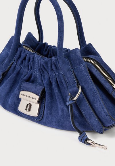 Sinine sametist käekott kogutud disainiga, hõbedaste detailide, tõmblukuga sulgemise ja kahekordsete käepidemetega. Esiküljel asub silmapaistev logoga plaat.