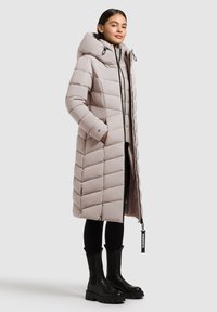 Podložena, svetlo bež dolga bunda s kapuco. Ima zadrgo na sprednji strani, vzorec v obliki črke V in črne detajle. V kombinaciji z črnimi gležnjarji.