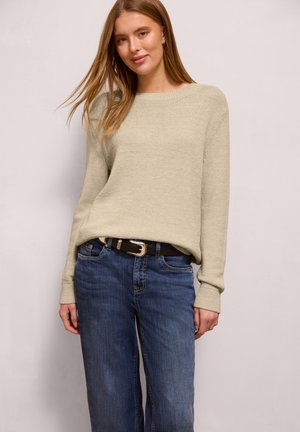 Ung kvinde med langt hår iført beige striktrøje og blå jeans med sort bælte, stående foran en ensfarvet lys baggrund.