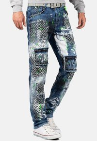 Jeans en denim présentant un look usé avec des éclaboussures de peinture verte, des textures contrastées, plusieurs poches et des clous en métal.