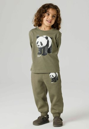 Child with curly hair wearing olive-green sweatshirt and pants featuring block-style panda graphics, standing with hands behind back.