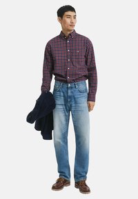 Mann trägt ein rot und marineblau kariertes Hemd, in hellblaue Jeans gesteckt, hält eine marineblaue Jacke und trägt braune Lederschuhe.