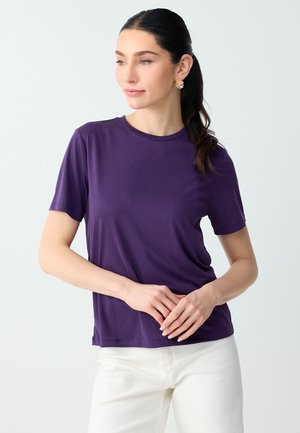 Donna con capelli scuri raccolti in una coda di cavallo, che indossa orecchini colorati, una maglietta viola a maniche corte e pantaloni bianchi su uno sfondo bianco uniforme.