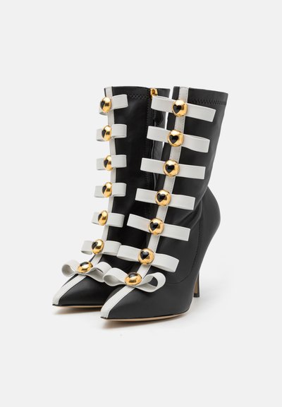 Botines de cuero negro con tiras horizontales blancas, acentos dorados, punteras afiladas y lazos decorativos en la parte frontal. Diseño de tacón alto.