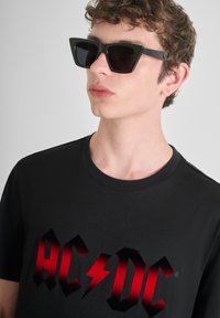 Camiseta negra con un gran logo rojo de AC/DC, combinada con gafas de sol negras angulares que presentan un acabado mate y lentes oscuros.