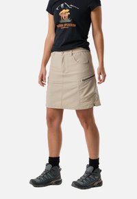 Kvinde iført sort t-shirt med grafik, beige udendørs nederdel med sidelomme med lynlås, sorte strømper og grå vandrestøvler, stående mod en hvid baggrund.