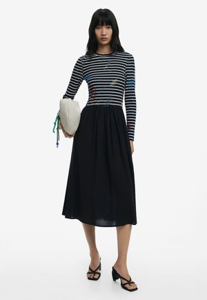 Striped long-sleeve top with colorful embroidery, black mid-length skirt, paired with a light clutch bag and strappy black heels.