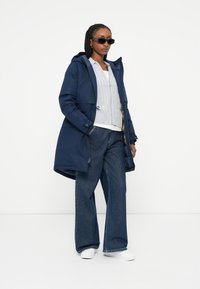 Parka blu navy con cappuccio, cerniere frontali e cordini, indossata sopra un maglione grigio a trecce e jeans a gamba larga, abbinata a sneakers bianche.