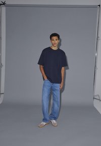 Mornarsko modra široka majica, svetlo modri ravni jeans in sivi športni copati. Model stoji proti sivi kulisi z nevtralno osvetlitvijo.