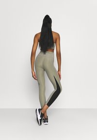 Žena s dlouhými vlasy, oblečená v šedých a černých legínách a sportovní podprsence, stojící s jednou nohou zkříženou za druhou na jednobarevném pozadí.