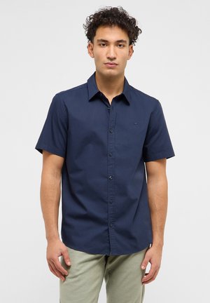 Junger Mann mit lockigem Haar, der ein marineblaues, kurzärmeliges Hemd mit Knöpfen und hellgraue Hose trägt, vor einem schlichten Hintergrund stehend.