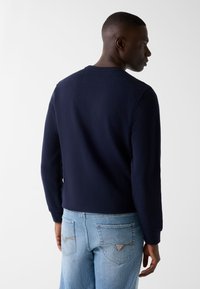 Sweat-shirt bleu marine à manches longues, col rond et ourlet côtelé. Porté avec un jean denim bleu clair avec des détails sur la poche arrière.