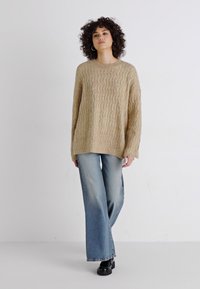 Beige strikkegenser med en teksturert, kabelmønster, sammen med lyseblå vide jeans og svarte sko. Modellen står mot en nøytral bakgrunn.
