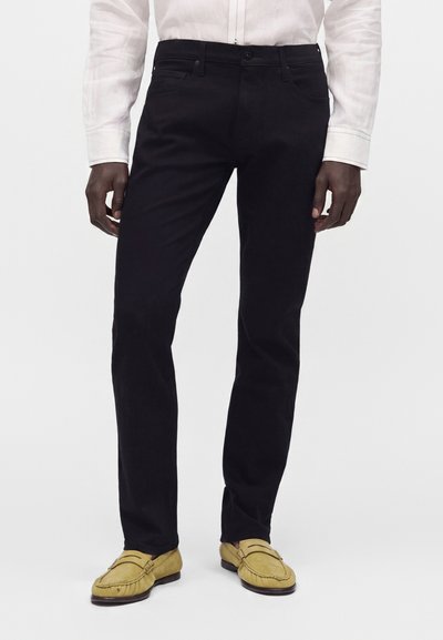 Άνδρας που φοράει μαύρο παντελόνι slim-fit, λευκό πουκάμισο με μακριά μανίκια και κίτρινες σουέντ μοκασίνια, στέκεται μπροστά σε απλό φόντο.