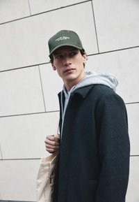 Grüner Baseballcap mit Logo, grauer Hoodie und dunkler Wollmantel. Hält eine beigefarbene Tragetasche; glatte Textur und strukturiertes Design.