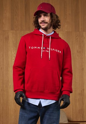 Hanorac roșu cu logo alb "TOMMY HILFIGER", cu cordon, buzunar frontal, combinat cu o cămașă albastră deschisă și blugi negri, purtat cu mănuși negre.