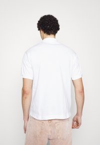 T-shirt polo blanc avec un col classique et des manches courtes, présenté de dos, associé à un pantalon en velours côtelé clair.