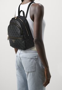 Mujer vestida con jeans de color azul claro, top blanco sin mangas y una mochila negra con patrón y cremalleras doradas en la espalda.