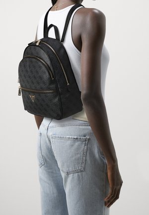 Kvinde iført lyseblå jeans, hvid ærmeløs top og en sort mønstret rygsæk med guldfarvede lynlåse på ryggen.