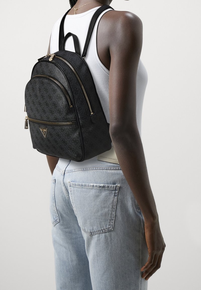 Mujer vestida con jeans de color azul claro, top blanco sin mangas y una mochila negra con patrón y cremalleras doradas en la espalda.