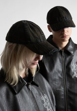 Două persoane purtând șepci negre de baseball uzate și jachete de piele negre cu broderii gri deschis, cu fața în jos pe un fundal simplu.