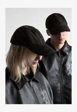 Deux personnes portant des casquettes de baseball noires usées et des vestes en cuir noires avec une broderie grise claire, regardant vers le bas sur un fond uni.